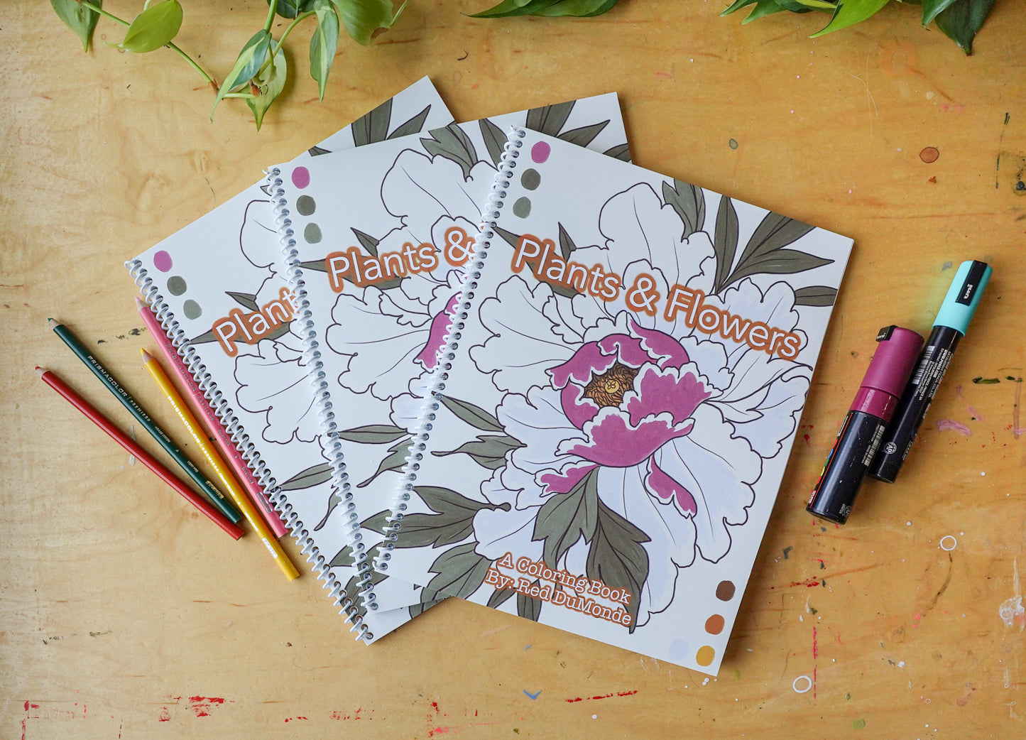 3 plants and flowers coloring books arranged on a wood table with colored pencils and posca pens posed next to the books and some leaves at the top of the image