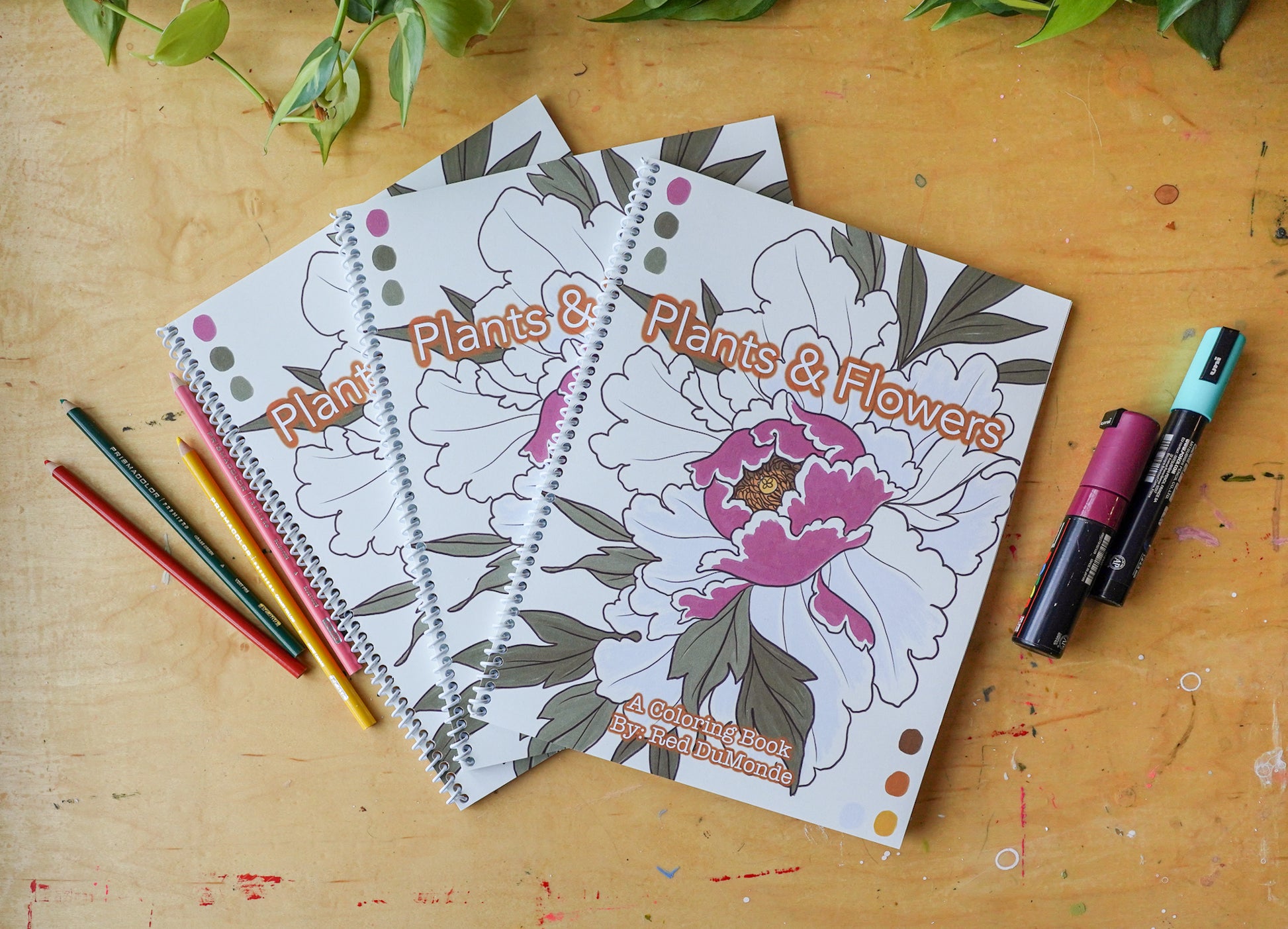 3 plants and flowers coloring books arranged on a wood table with colored pencils and posca pens posed next to the books and some leaves at the top of the image
