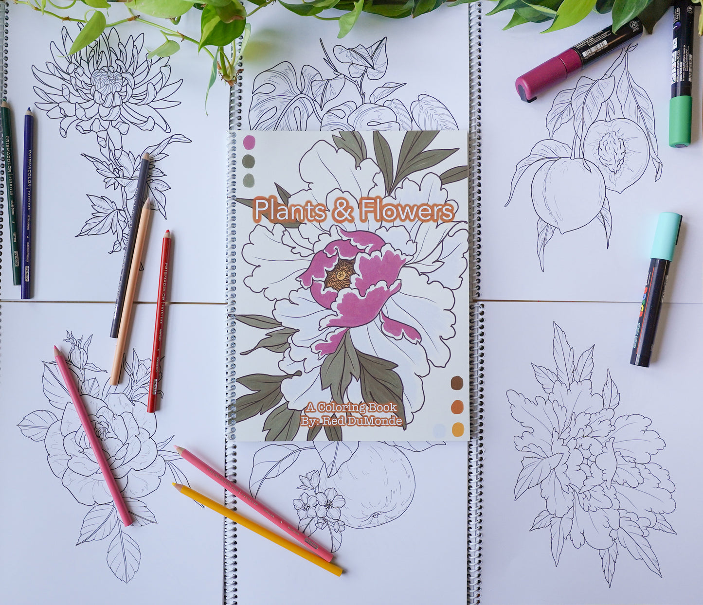 Coloring book laying on top of a grid of coloring book pages, with some colored pencils and posca pens scattered about and some leaves at the top of the image