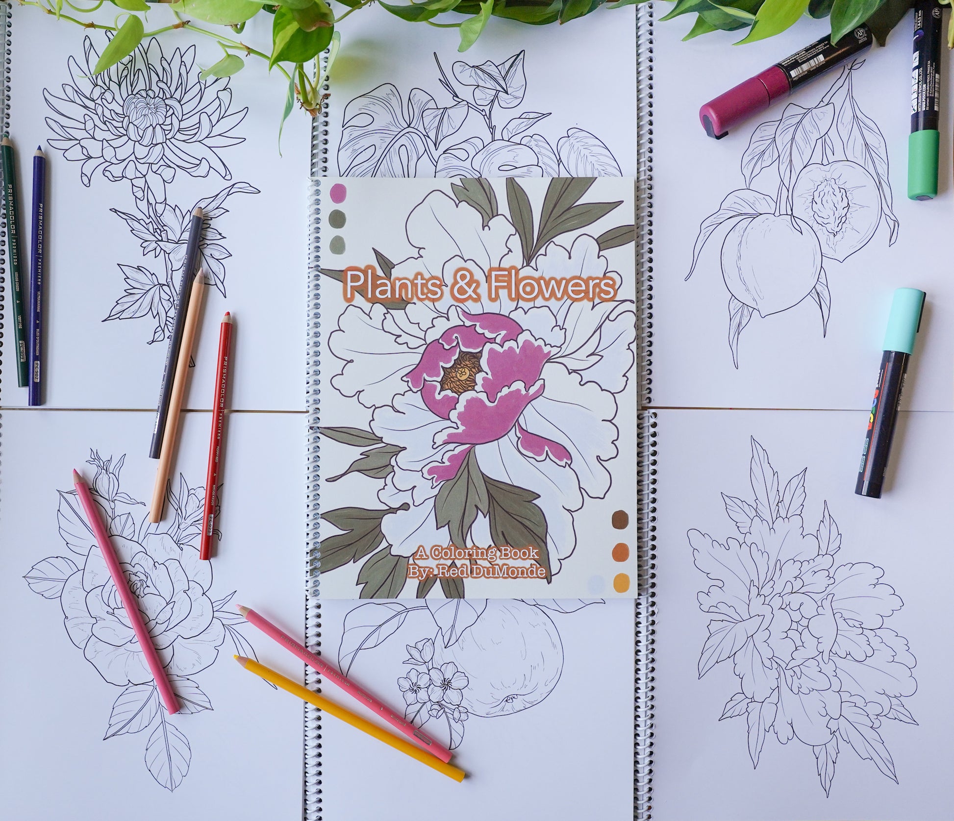 Coloring book laying on top of a grid of coloring book pages, with some colored pencils and posca pens scattered about and some leaves at the top of the image