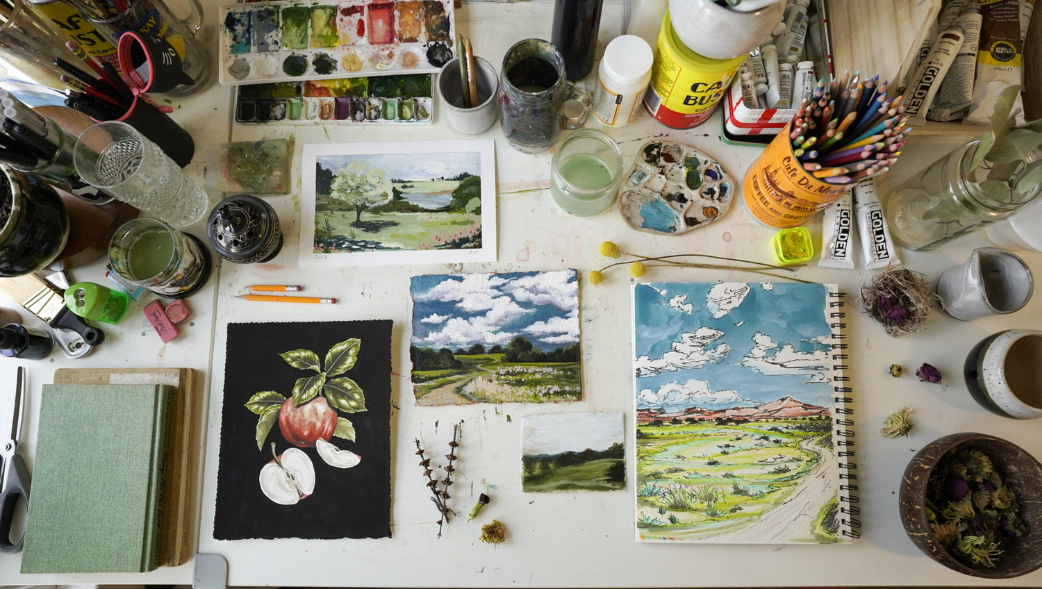 Nature inspired artwork including botanical illustrations and landscape paintings displayed in a flat lay on a studio work table with art supplies
