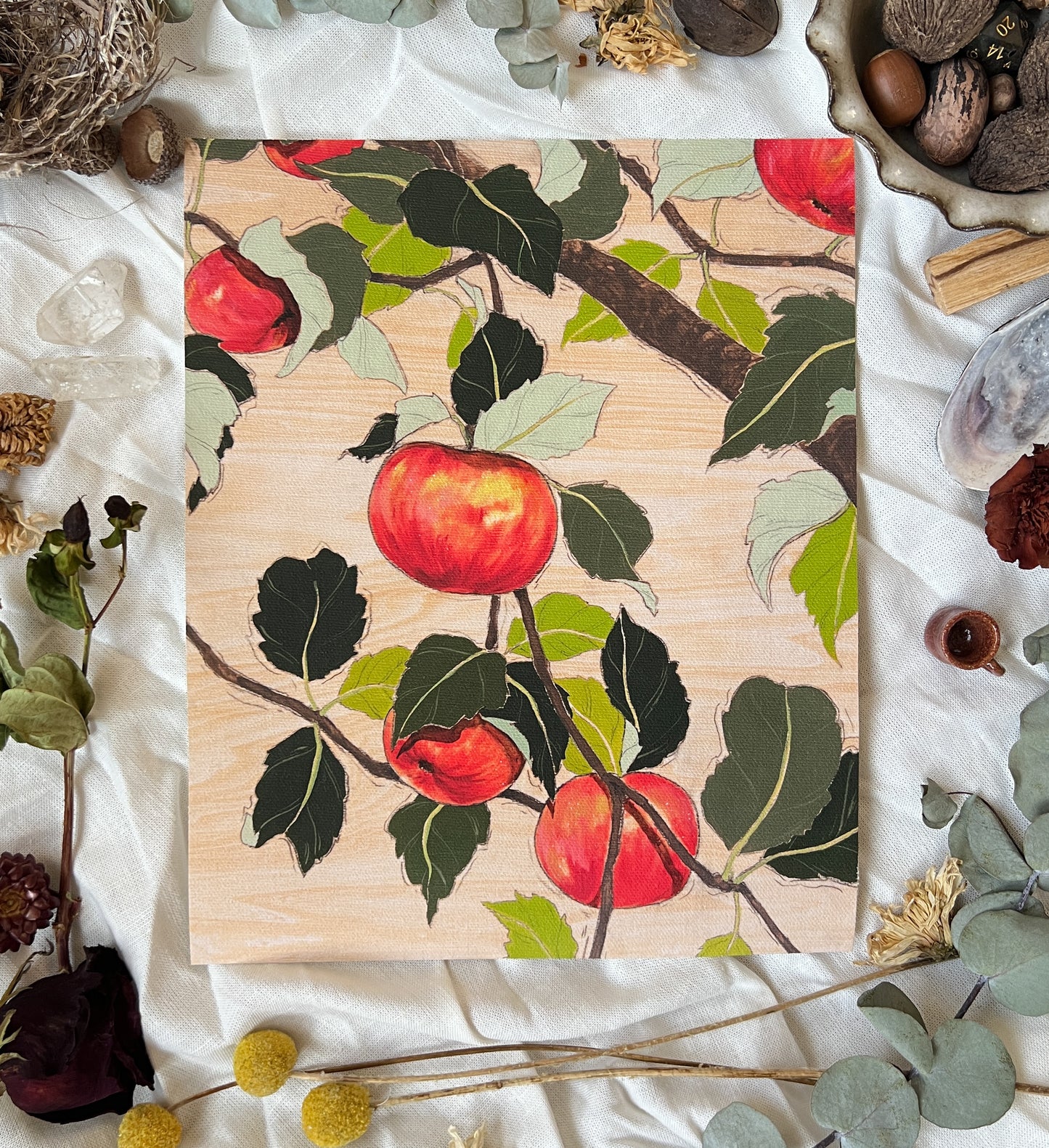 Art print of red apples on branches with wood grain background. Print is sitting on a white linen sheet surrounded by dried plants and other nature items.