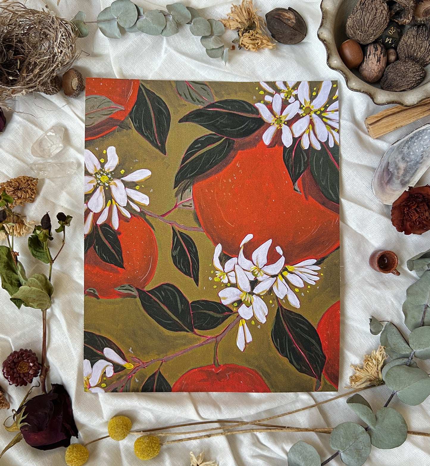 Art print of Oranges and orange blossoms on branches with green background. Print is sitting on a white linen sheet surrounded by dried plants and other nature items.