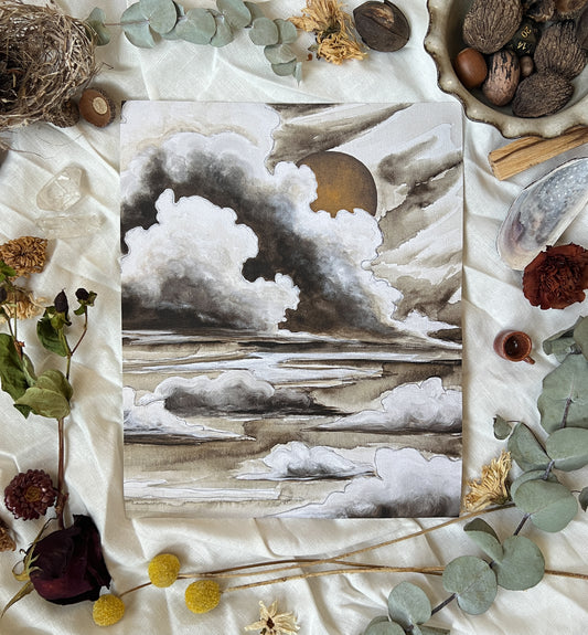 Art print of moody, neutral colored clouds with a dark gold sun. Print is sitting on a white linen sheet surrounded by dried plants and other nature items.