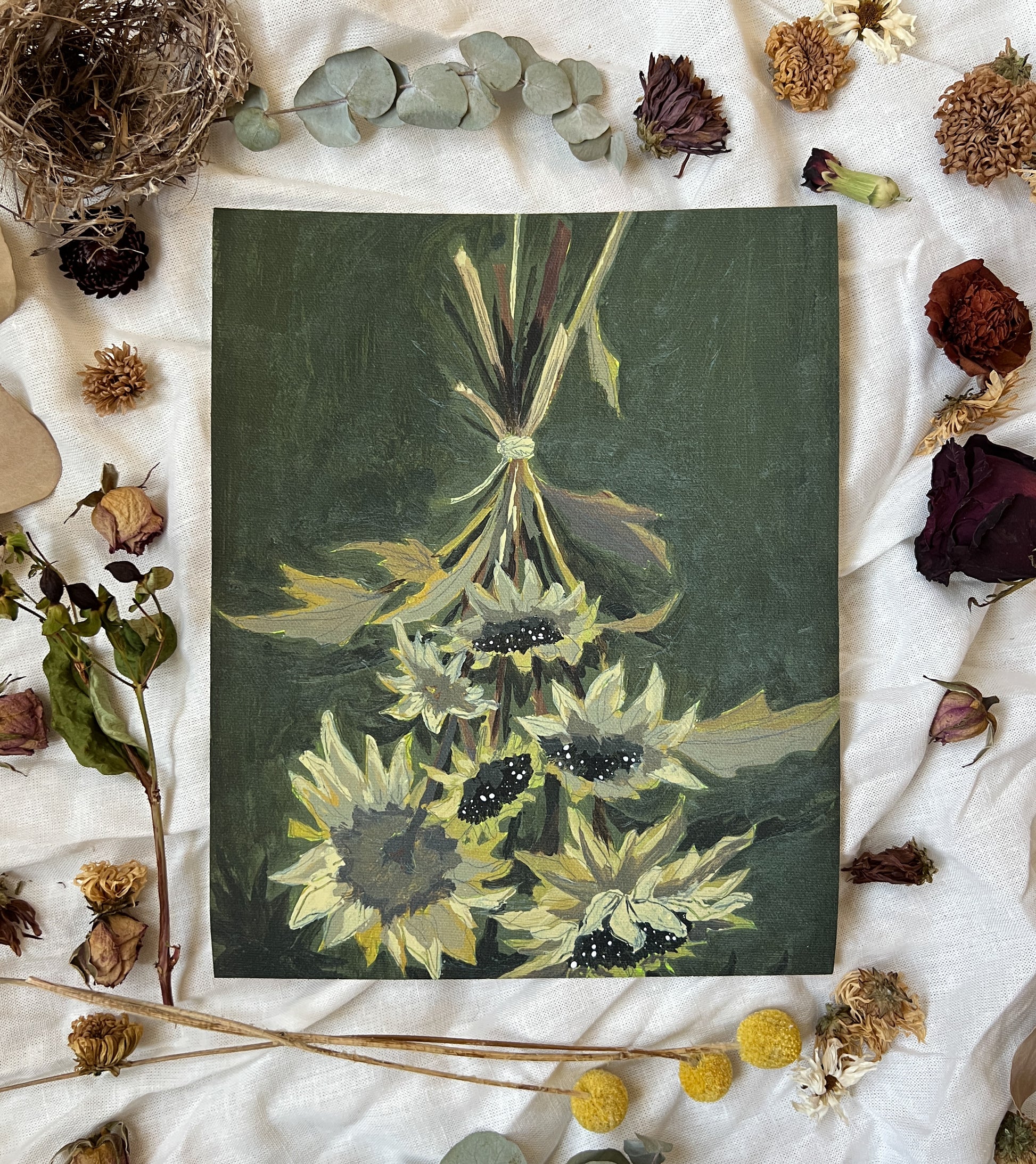 Art print of dried sunflowers with dark green background. Print is on a white linen cloth with natural dried flowers and other nature items surrounding it.