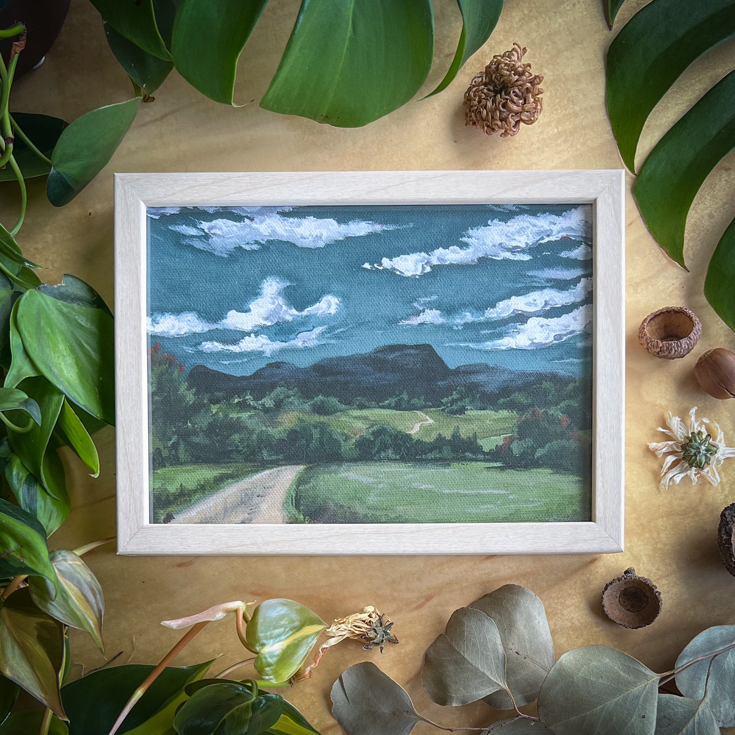 Canvas print of a mountain landscape in a light wood frame displayed on a wood table with plants and other natural items scattered around it