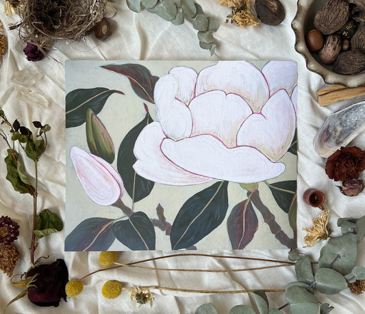 Art print of magnolia blossom and buds on branches with neutral background. Print is sitting on a white linen sheet surrounded by dried plants and other nature items.