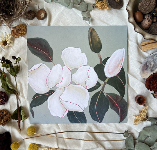 Art print of magnolia blossom and buds on branches with grey/blue background. Print is sitting on a white linen sheet surrounded by dried plants and other nature items.