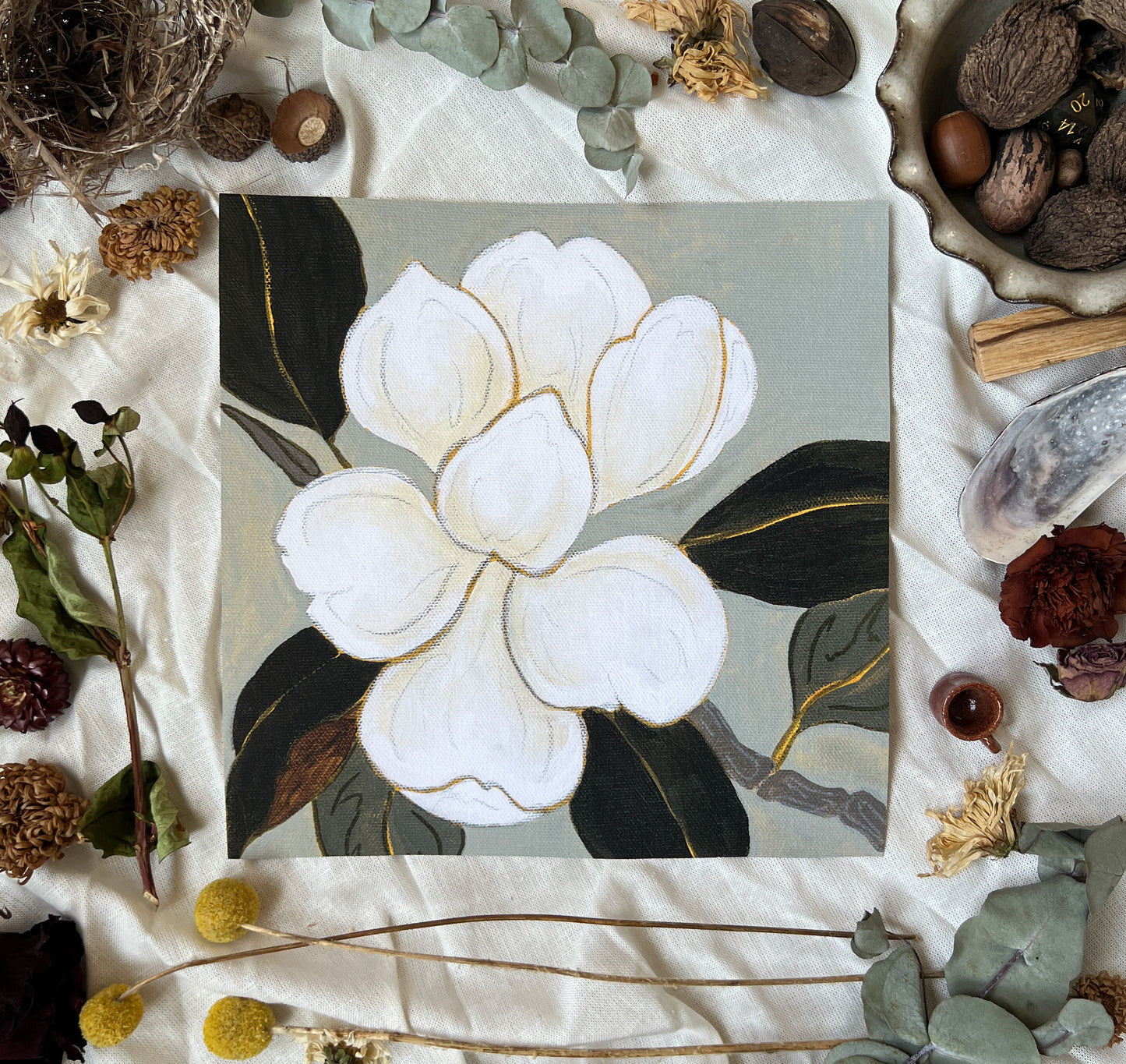 Art print of magnolia bloom on branch with grey/blue background. Print is sitting on a white linen sheet surrounded by dried plants and other nature items.