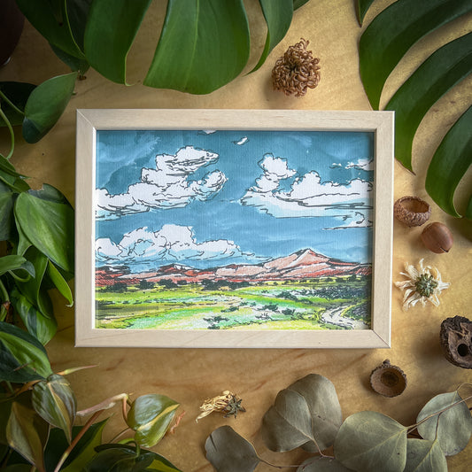 Canvas print of a new mexico landscape with puffy clouds and pink mountains in a light wood frame displayed on a wood table with plants and other natural items scattered around it