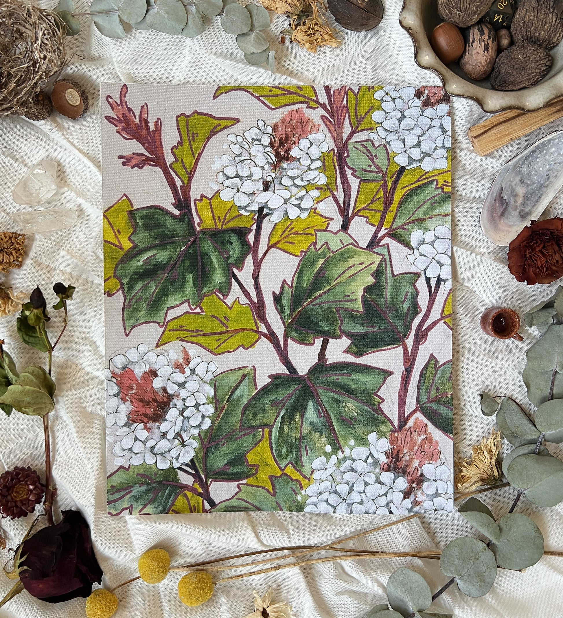 Art print of white oak leaf hydrangea bush with canvas color background. Print is sitting on a white linen sheet surrounded by dried plants and other nature items.