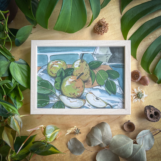 Canvas print of a pear still life in a light wood frame displayed on a wood table with plants and other natural items scattered around it
