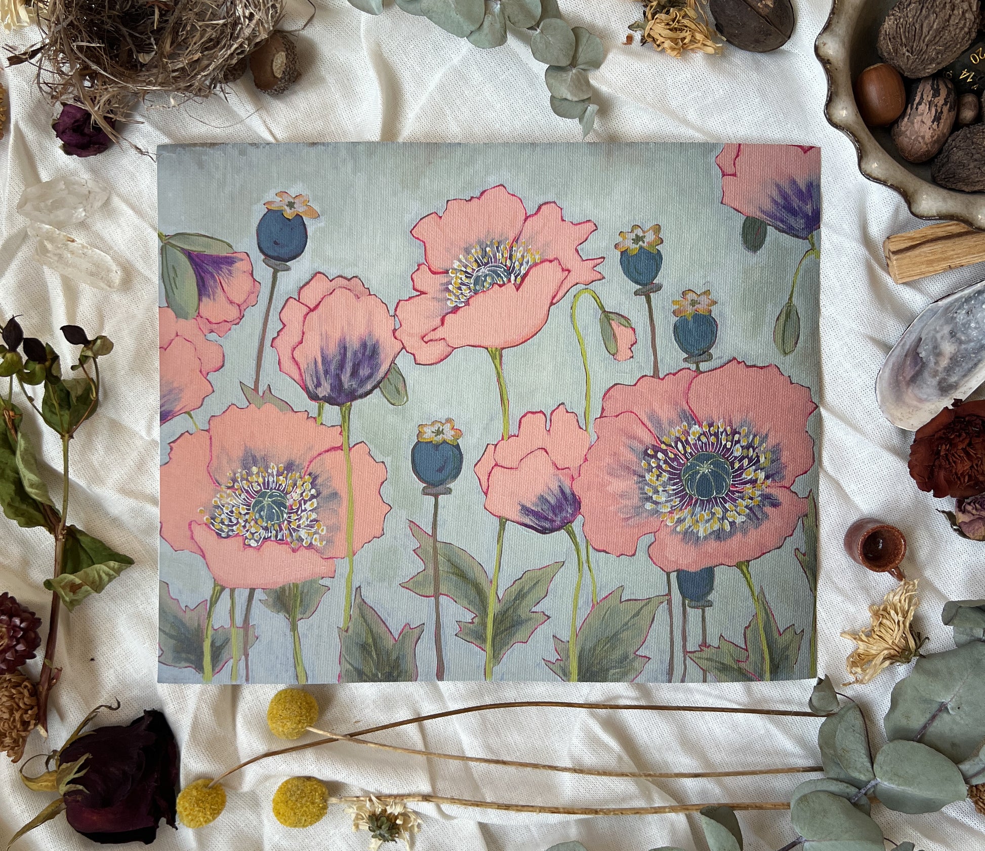 Art print of pink poppy flowers with a grey/blue background. Print is sitting on a white linen sheet surrounded by dried plants and other nature items.