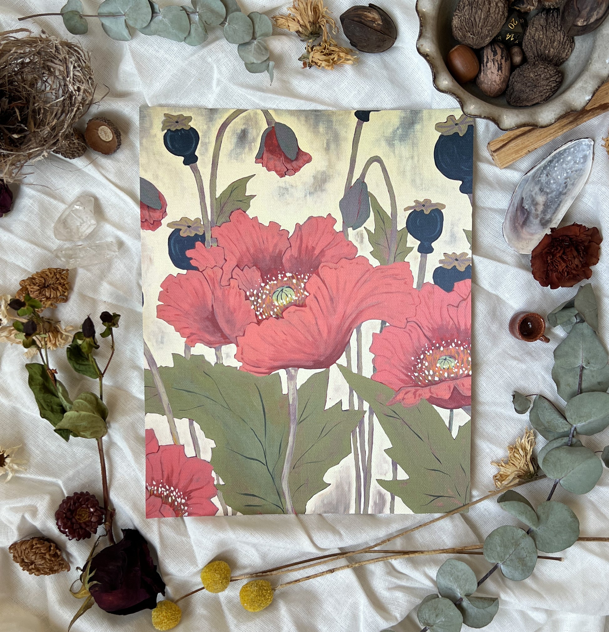 Art print of pink poppies with buds and seed pods on a neutral background. Print is sitting on a white linen sheet surrounded by dried plants and other nature items.