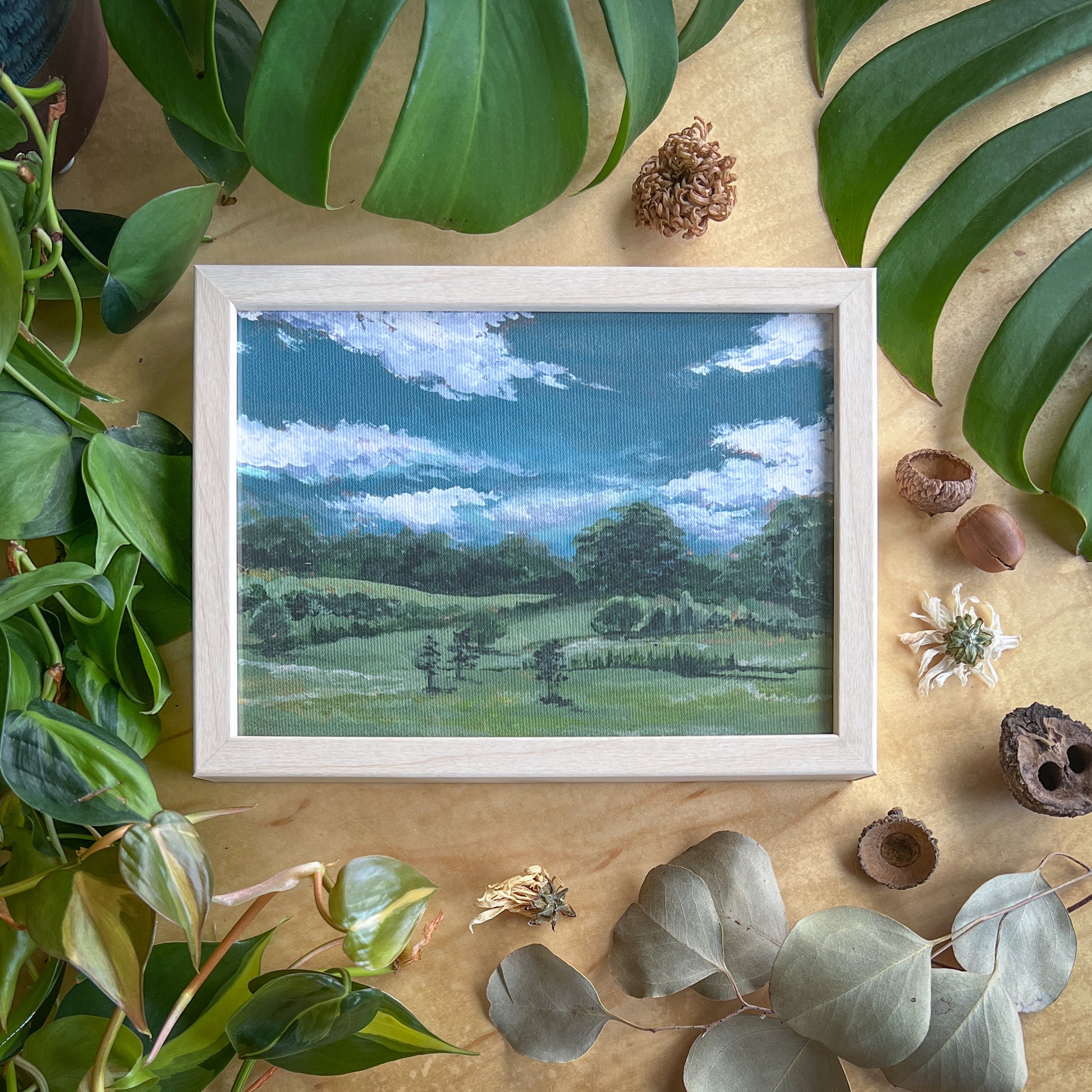 Canvas print of a landscape in a light wood frame displayed on a wood table with plants and other natural items scattered around it