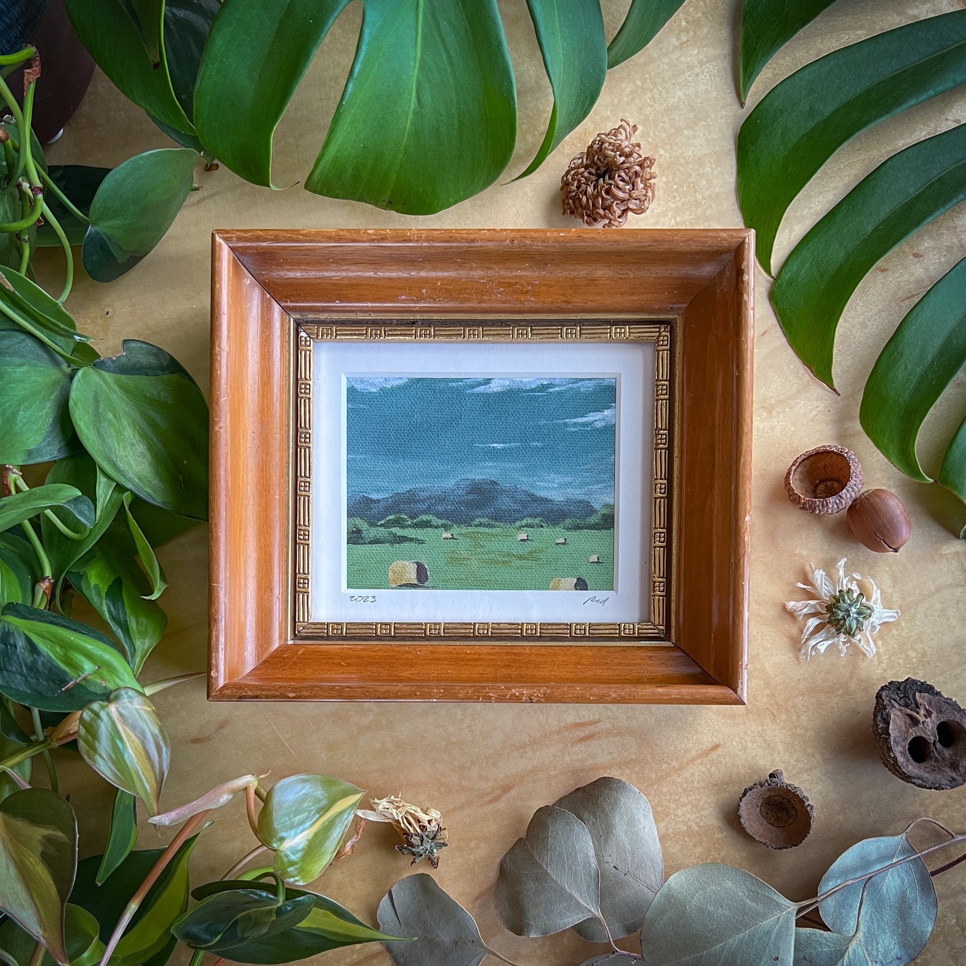 Canvas print of a landscape in a wood frame displayed on a wood table with plants and other natural items scattered around it