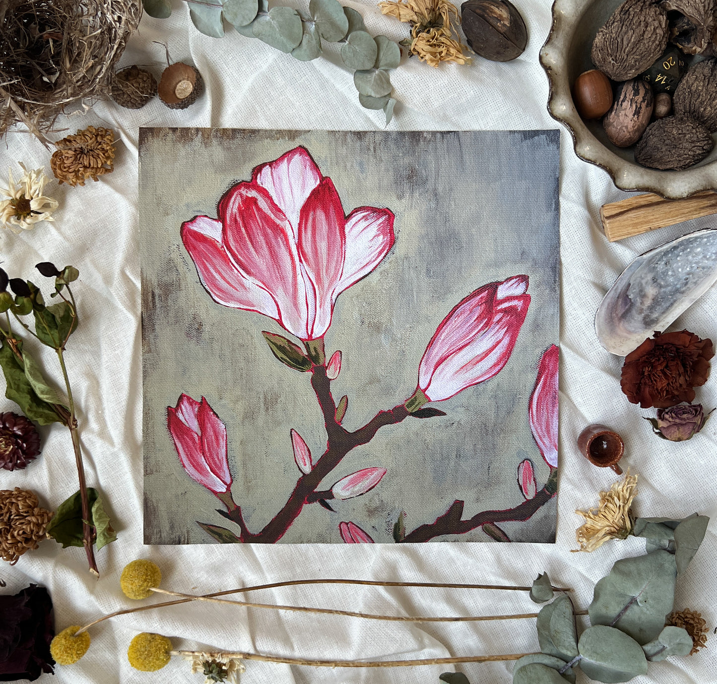 Art print of pink and white Tulip magnolia flowers on branches. Print is sitting on a white linen sheet surrounded by dried plants and other nature items.