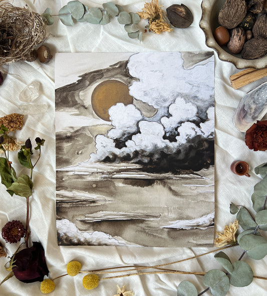 Art print of moody, neutral colored clouds with a dark gold sun. Print is sitting on a white linen sheet surrounded by dried plants and other nature items.